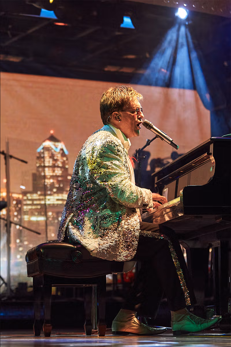 A photo of a Crystal Cruises performer dressed as a famous musician (either Elton John or Billy Joel) on a stage with spotlights and concert lighting. The performer is singing into a microphone and engaging with the audience. (Photo Courtesy Crystal Cruises)