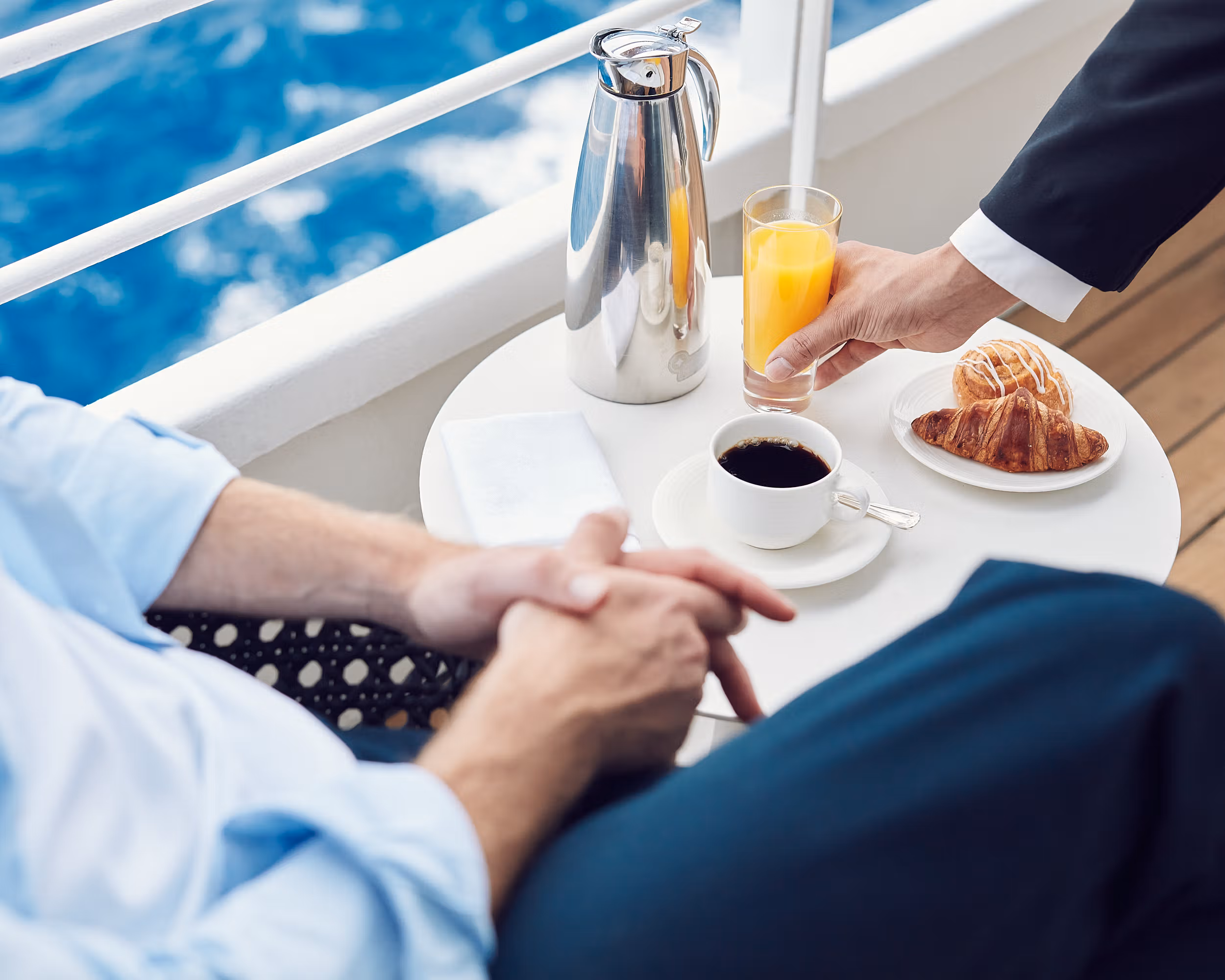 A photo of a Crystal Serenity suite veranda with a breakfast table. The table features a white tablecloth, pastries, coffee, and orange juice. Comfortable seating allows guests to enjoy their breakfast while gazing out at the ocean on their in-suite veranda. (Photo Courtesy Crystal Cruises)